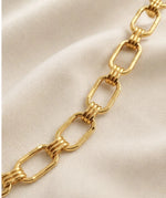 A close-up of a gold chain link bracelet with elongated oval links and twisted ring connectors, laid diagonally on a soft beige fabric background.