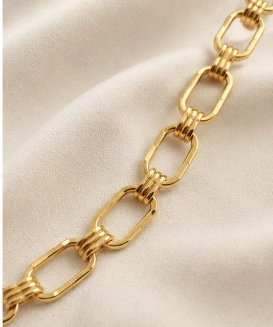 A close-up of a gold chain link bracelet with elongated oval links and twisted ring connectors, laid diagonally on a soft beige fabric background.